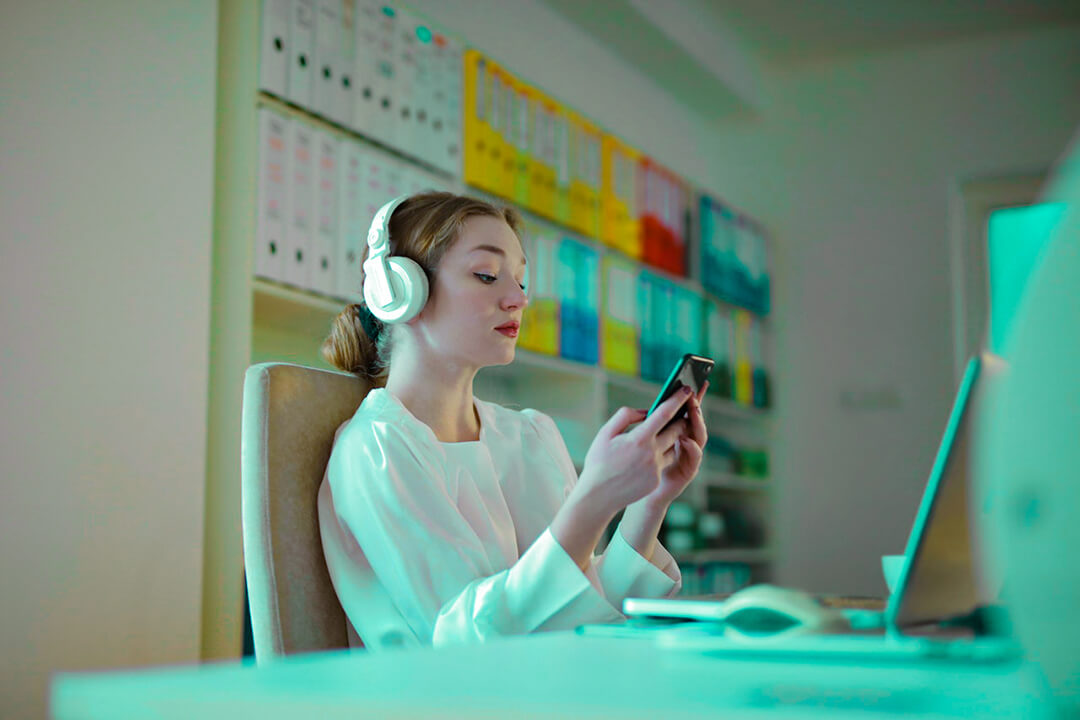 woman with headset on in front of her desktop computer browsing through her smartphone